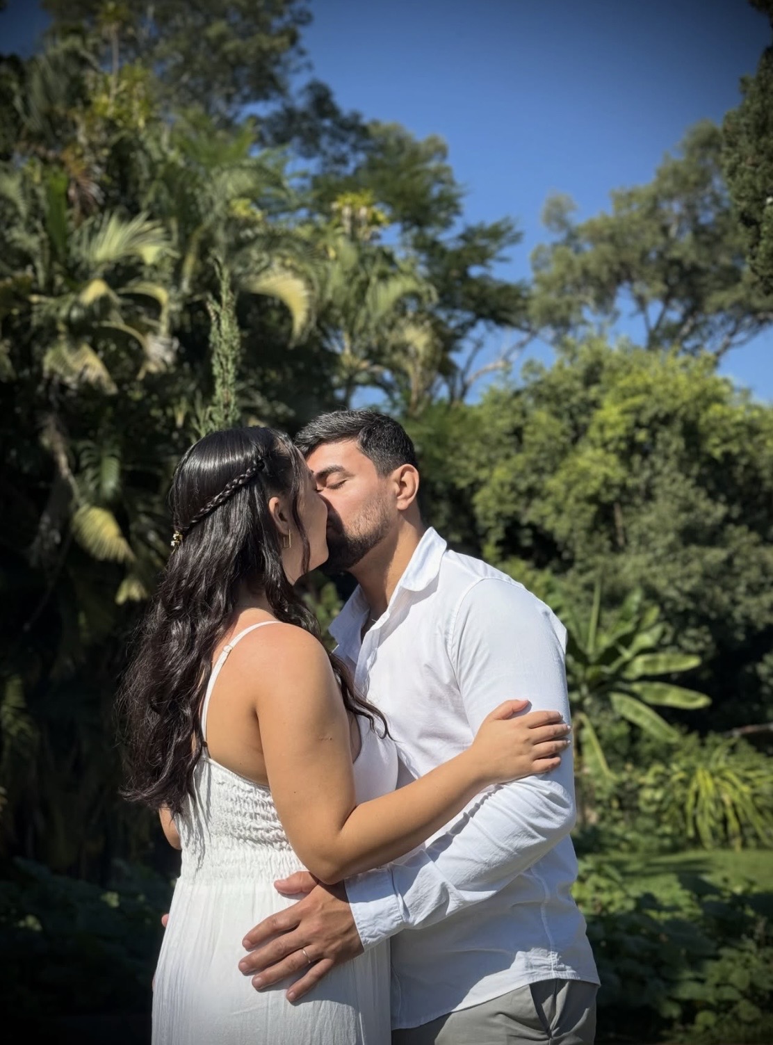 Casal se beijando em jardim ensolarado, ambos de branco