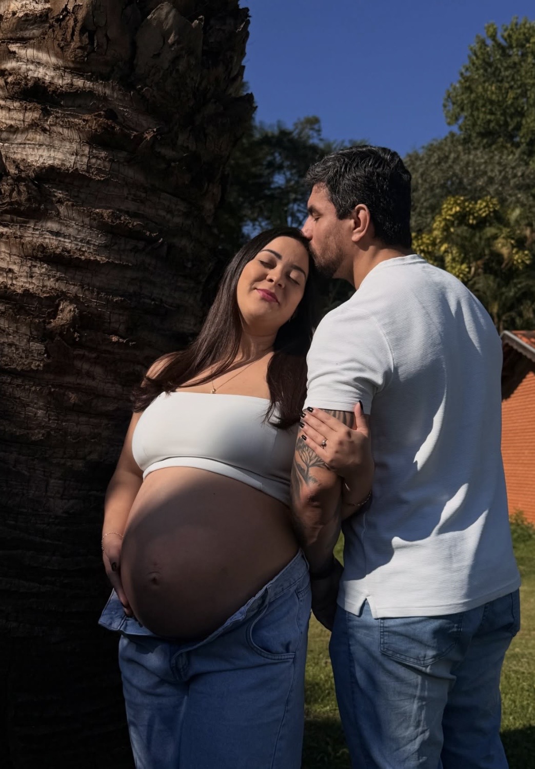 Casal em ensaio de gestante junto a uma palmeira, ela de top branco e jeans